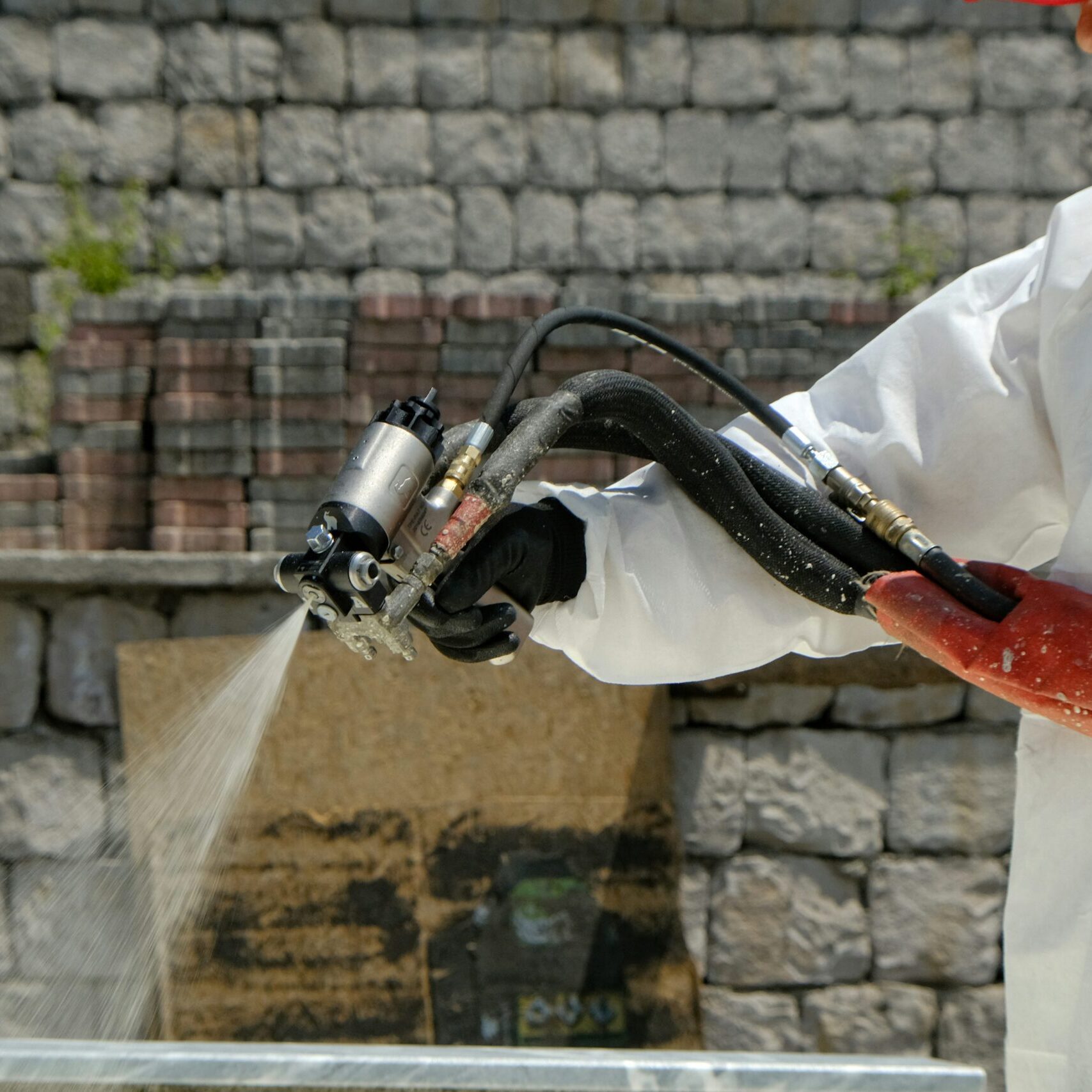 A worker in a white protective suit and red gloves uses a handheld sprayer to spray liquid on a surface.